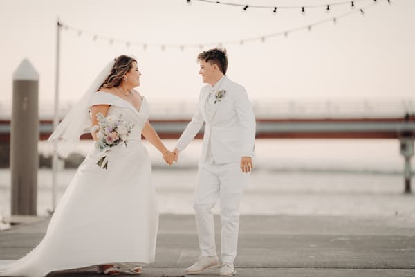 Brooke and Tiffany hold hands outdoors near water with a bridge and string lights in the background, both dressed in white wedding attire; Brooke wears a wedding gown and veil holding a bouquet, Tiffany wears a white suit with a boutonniere.
