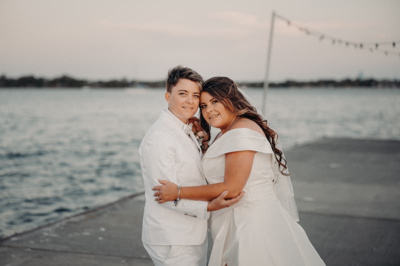 Brooke and Tiffany pose together near water at an outdoor location, with Brooke wearing a white suit and Tiffany in an off-shoulder white wedding dress with a veil.