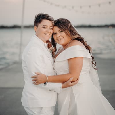 Brooke and Tiffany pose together outdoors near water, with Brooke wearing a white suit and Tiffany in an off-shoulder white wedding gown at Sandstone Point Hotel.