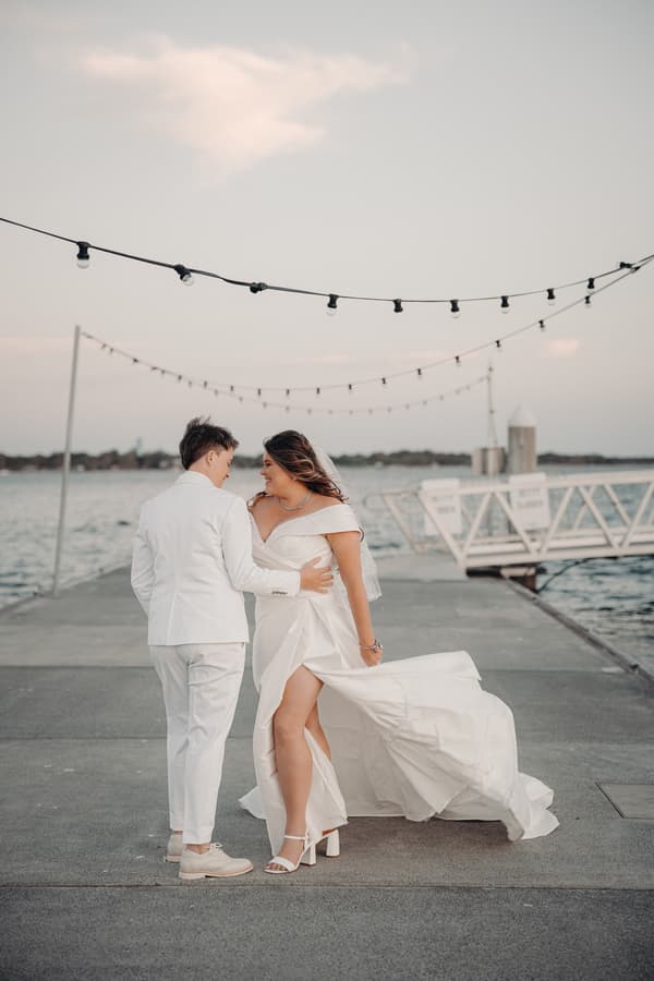 Brooke and Tiffany stand together on a pier by the water, with string lights overhead. Brooke wears a white off-shoulder wedding dress with a slit, and Tiffany wears a white suit. They face each other, holding each other closely.