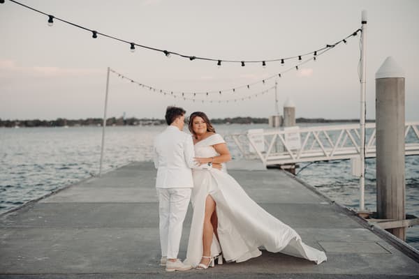 Brooke and Tiffany pose together on a jetty, with water and string lights visible in the background. Engagement ceremony by She Said Yes.