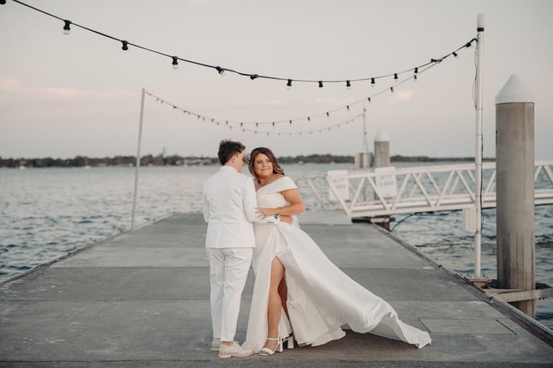 Brooke and Tiffany pose together on a jetty at Sandstone Point Hotel, with water and string lights visible in the background.