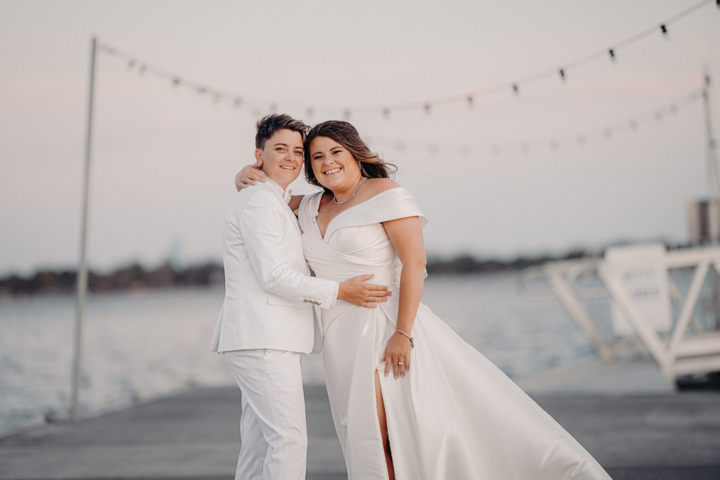 Brooke and Tiffany pose together on a pier with water and string lights in the background. Engagment party by She Said Yes.