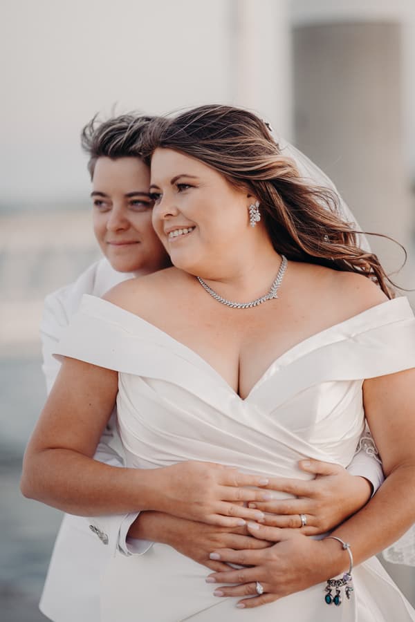 Brooke and Tiffany embrace outdoors in a styled couple portrait, with Brooke wearing an off-shoulder wedding dress and Tiffany in a white suit.