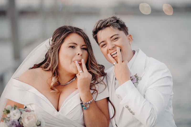 Brooke and Tiffany pose together outdoors, both showing their wedding rings with their fingers near their mouths. Brooke wears an off-shoulder wedding dress and veil, holding a bouquet, while Tiffany wears a white suit with a boutonniere.