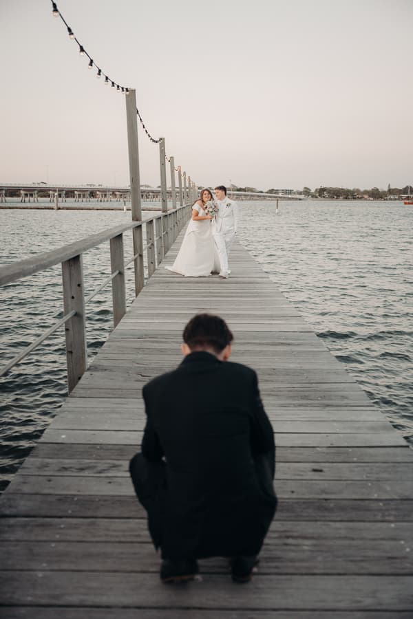 Brooke and Tiffany pose together on a wooden pier over water at Sandstone Point Hotel for a styled couple portrait while a photographer crouches in the foreground.