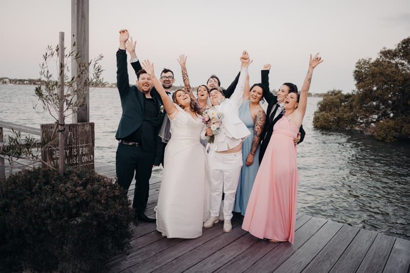 Brooke and Tiffany celebrate with their bridal party on a wooden jetty by the water at Sandstone Point Hotel — The Pavilion.