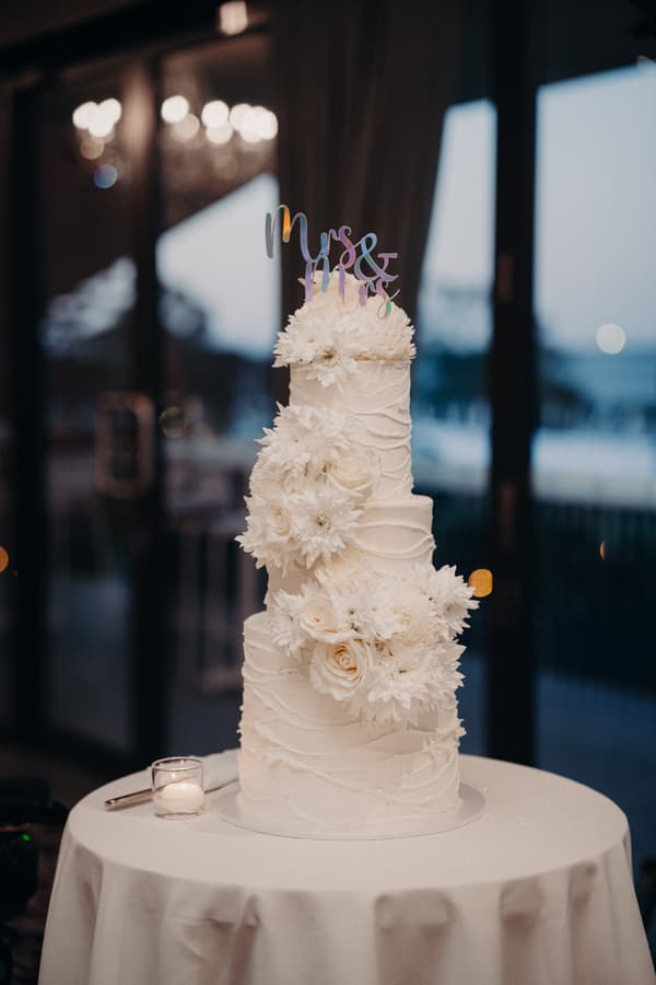 A three-tier white wedding cake decorated with white flowers and a topper reading 'Mrs & Mrs' on a round table at Sandstone Point Hotel — The Pumicestone Room.