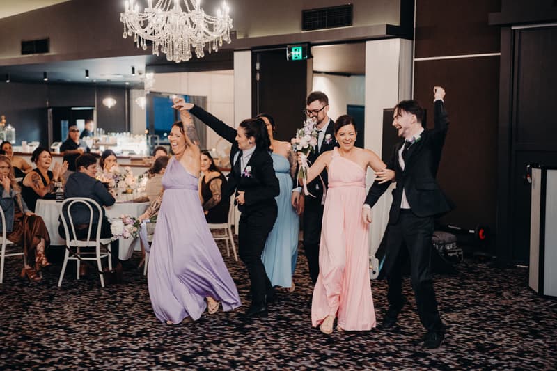 Bridal party members dance and celebrate on the reception stage at Sandstone Point Hotel — The Pumicestone Room, with guests seated at tables in the background.