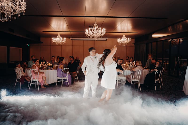 Brides Brooke and Tiffany dance on a fog-covered dance floor at their wedding reception in The Pumicestone Room at Sandstone Point Hotel, while guests seated at round tables watch.
