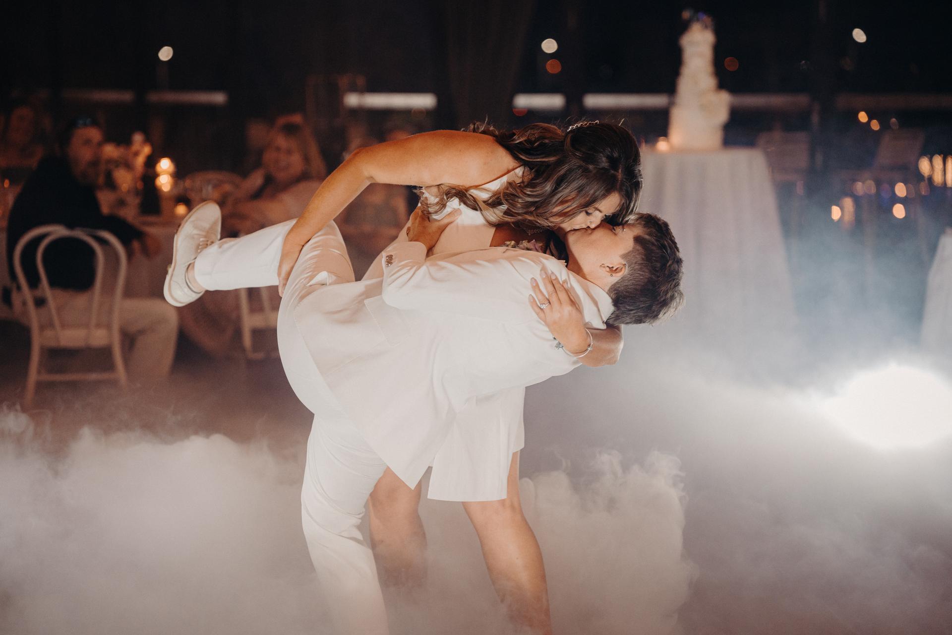 Brooke and Tiffany share a kiss during their first dance at the reception in The Pumicestone Room, Sandstone Point Hotel, with guests seated at tables in the background and a wedding cake visible on a table.