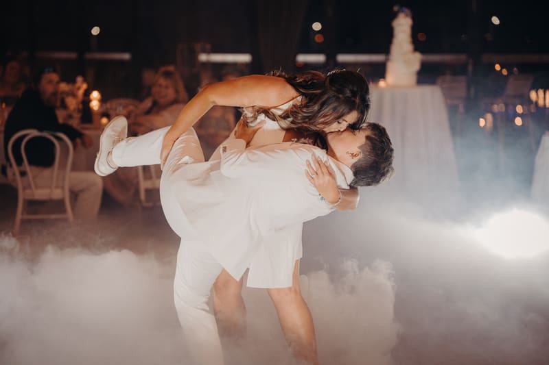 Brooke and Tiffany share a kiss during their first dance at the reception in The Pumicestone Room, the venue, with guests seated at tables in the background and a wedding cake visible on a table. Cinematic videographer by She Said Yes.