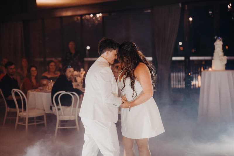 Brooke and Tiffany share a dance on the reception stage at Sandstone Point Hotel — The Pumicestone Room, with guests seated at tables in the background and a wedding cake on a table to the right.