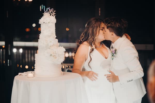 Brides Brooke and Tiffany kiss beside a white tiered wedding cake decorated with flowers and a "Mrs & Mrs" topper at Sandstone Point Hotel — The Pumicestone Room.