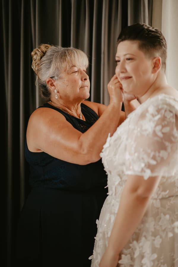 An older woman adjusts the earring of a bride wearing a white floral lace wedding dress in front of dark curtains.