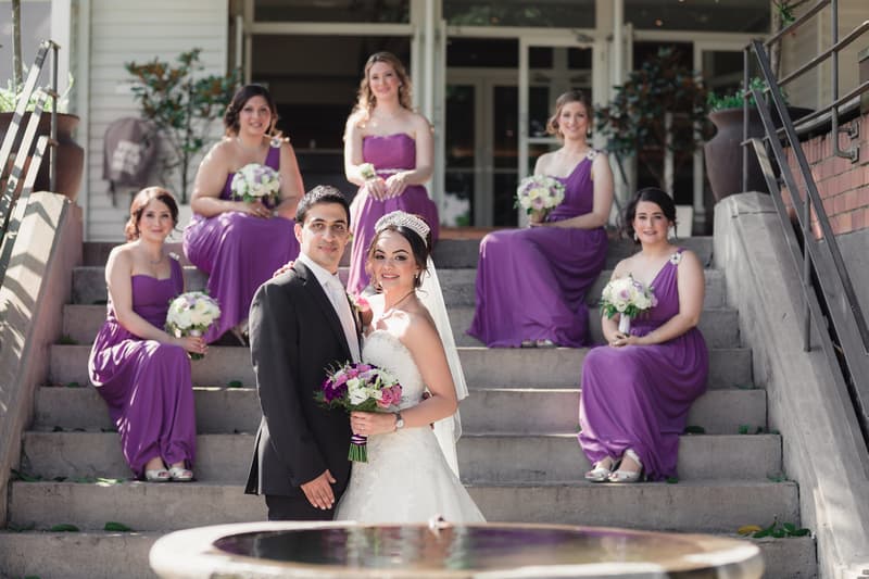 The bride and groom stand together on steps outside Hillstone St Lucia — The Quartyard, with five bridesmaids in purple dresses seated behind them holding bouquets.