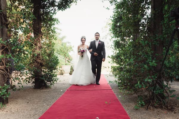 The bride Maryam and the groom Pasha walk arm-in-arm down a red carpeted outdoor aisle framed by greenery at Hillstone St Lucia — The Quartyard.