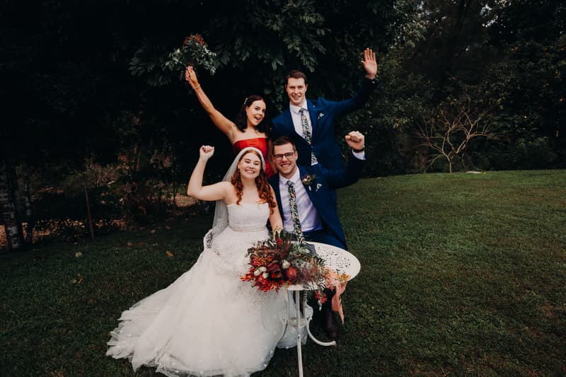 The bride Lilly in a white wedding gown and the groom Connor in a blue suit sit at a small white table with a floral arrangement at Yabbaloumba Retreat — By The River, accompanied by a bridesmaid in a red dress and a groomsman in a blue suit standing behind them, all raising their arms in celebration outdoors on grass with trees in the background.