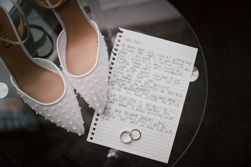 The bride's white pearl-embellished shoes, a handwritten wedding vow letter, and two wedding rings are displayed on a glass table at Sandstone Point Hotel.