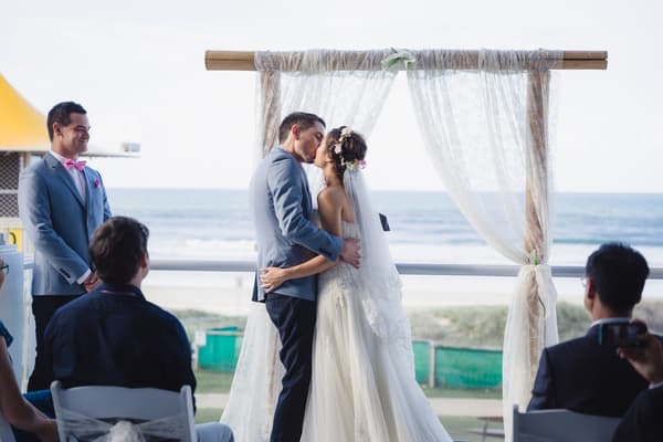 Bride Wing and groom Jason kiss at the altar during their wedding ceremony at Bilinga Beach Weddings — The Terrace, with a groomsman and seated guests watching.