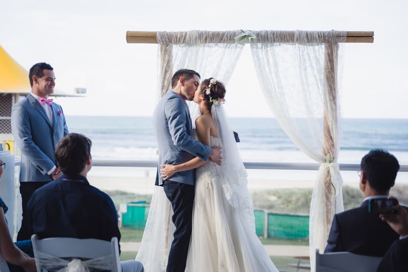 Bride Wing and groom Jason kiss at the altar during their wedding ceremony, with a groomsman and seated guests watching. Elopement photographer Brisbane by She Said Yes.