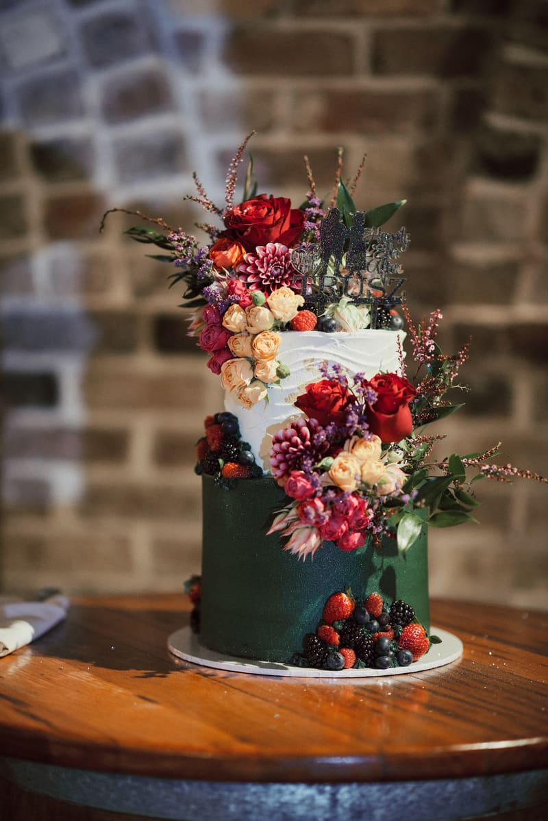 Two-tier wedding cake decorated with red and peach flowers and assorted berries, displayed on a wooden table. Valentine's Day wedding photography by She Said Yes.