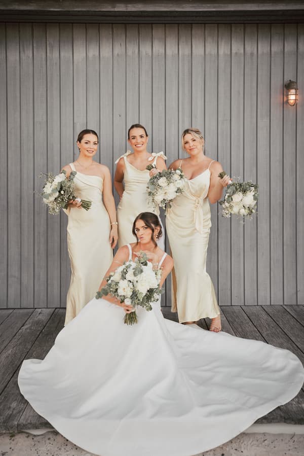 The bride Ashleigh poses seated on a wooden deck in front of a grey wooden wall at Sandstone Point Hotel, holding a bouquet of white flowers, with three bridesmaids standing behind her in matching champagne-colored dresses, each holding similar bouquets.