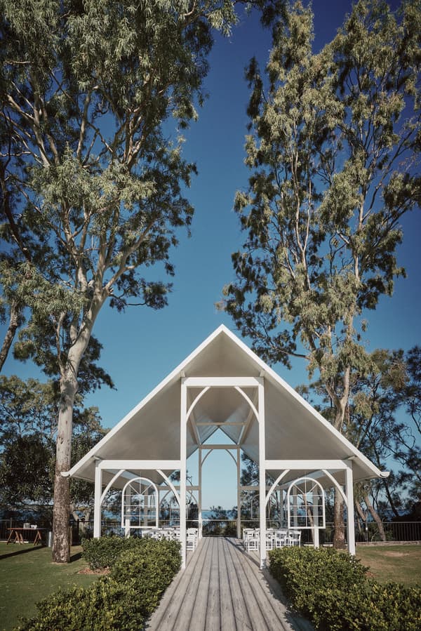Empty white pavilion structure with rows of white chairs set up for a wedding ceremony at Sandstone Point Hotel — The Pavilion, surrounded by tall trees and greenery under a clear blue sky.