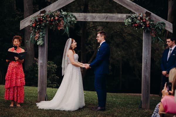 Bride Lilly and groom Connor hold hands facing each other under a wooden arch decorated with greenery and flowers at Yabbaloumba Retreat — By The River. An officiant in a red dress reads from a book to the left, and a groomsman in a blue suit stands to the right. Guests are partially visible in the foreground.