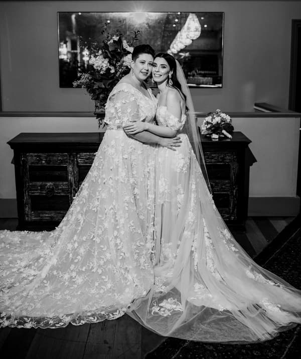 Two brides in wedding gowns embrace and smile at the camera indoors, standing in front of a wooden sideboard with floral arrangements and a large mirror above it.