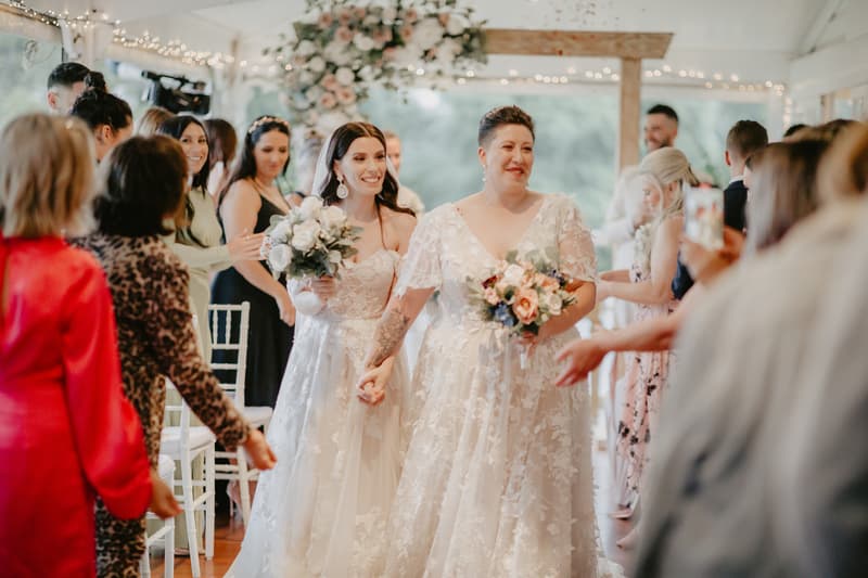 Two brides in white wedding dresses holding bouquets walk down an aisle lined with guests inside a decorated venue with string lights and floral arrangements.