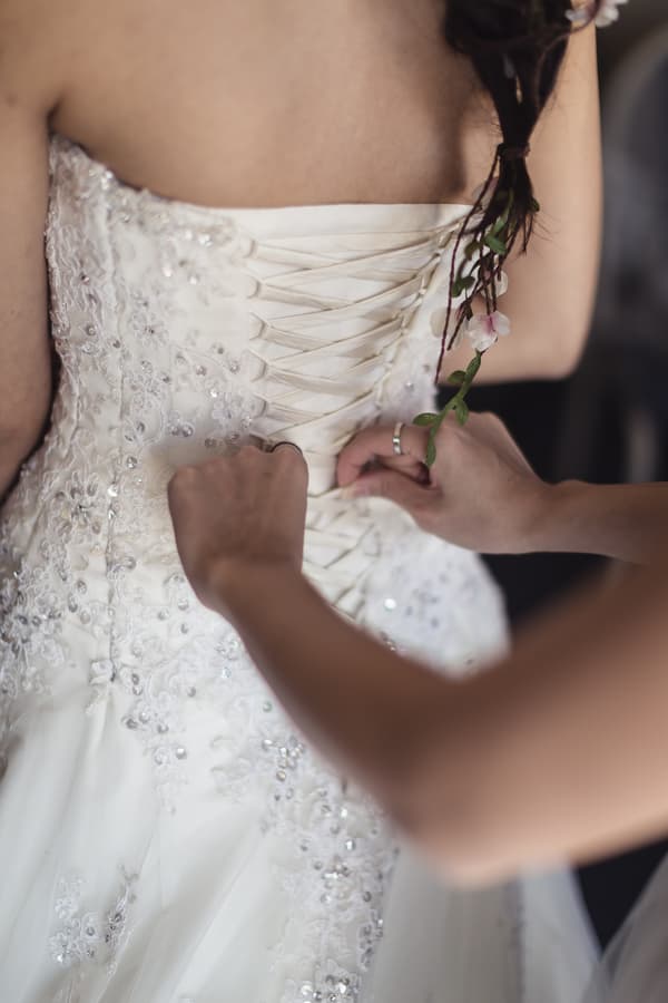 The bride is having the back of her wedding dress laced up by another person during preparation.