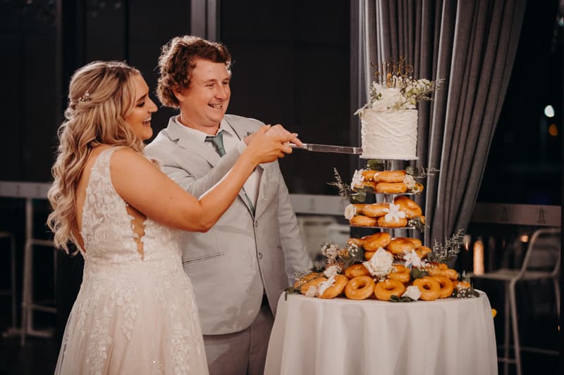 Bride Chloe and groom Brodie cut their wedding cake and donut tower at the Sandstone Point Hotel — Pumicestone Room during the reception.