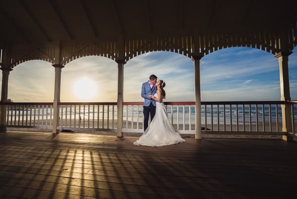 The bride Wing and groom Jason embrace and kiss under a wooden pavilion overlooking the beach at Bilinga Beach Weddings during sunset.