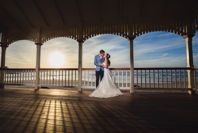 The bride Wing and groom Jason embrace and kiss under a wooden pavilion overlooking the beach at Bilinga Beach Weddings during sunset.