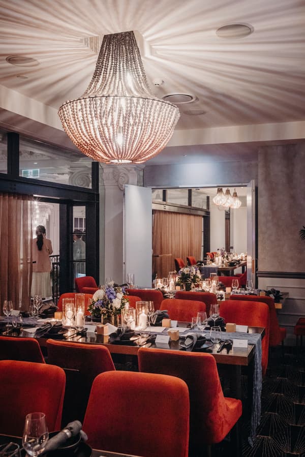 Reception dining area at Donna Chang with tables set with floral centerpieces, candles, glassware, and red chairs; two guests stand near the window in the background.