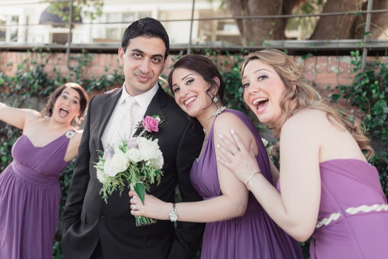 The groom Pasha poses with three bridesmaids in purple dresses at Hillstone St Lucia — The Quartyard, one bridesmaid holding a bouquet and another photobombing in the background.