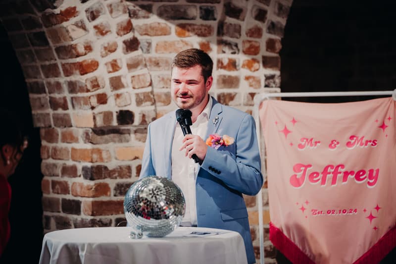A man in a light blue suit with a floral boutonniere holds a microphone and stands behind a round table with a disco ball on it at Sandstone Point Hotel — Cellar. A pink banner with the text 'Mr & Mrs Jeffrey Est. 02.10.24' is visible in the background.