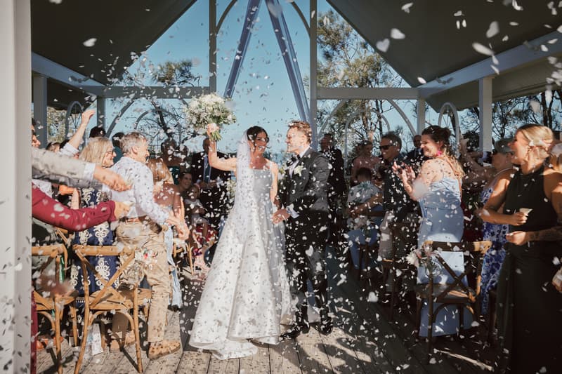 Bride Ashleigh and groom James walk down the aisle at Sandstone Point Hotel — The Pavilion as guests throw white confetti.