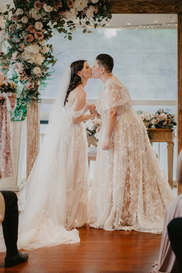 Two brides in lace wedding gowns hold hands and kiss under a floral arch indoors during a wedding ceremony.