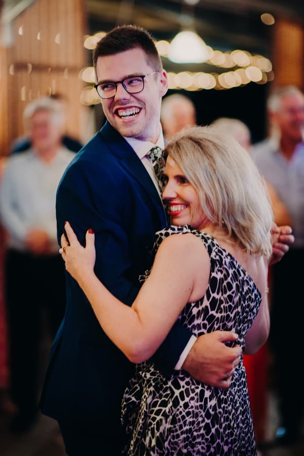 Connor in a dark suit and glasses dances with a woman in a patterned dress at the reception stage of Yabbaloumba Retreat — The Shed, with blurred guests and string lights in the background.