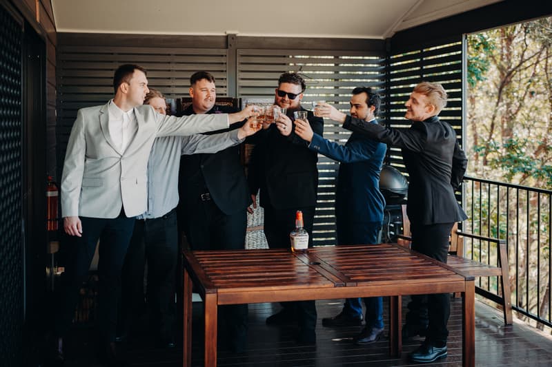 Adam and five groomsmen raise glasses in a toast on a covered balcony at Ocean View Estates.