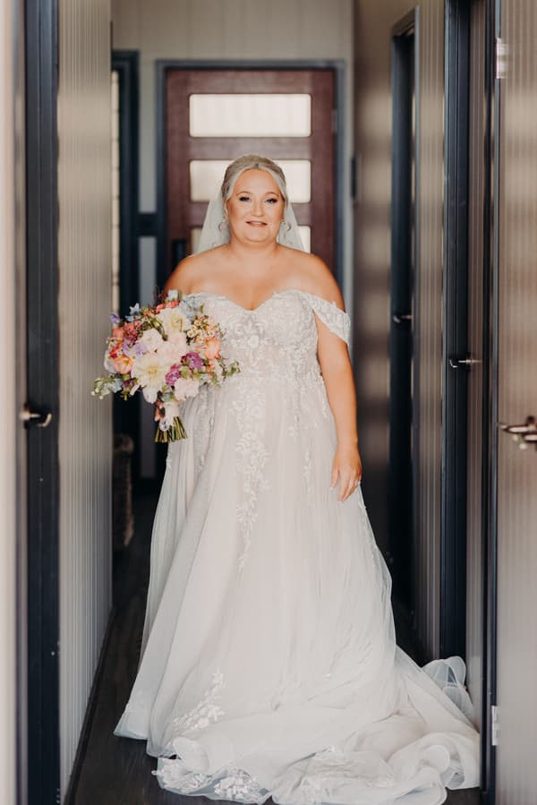 The bride Chantelle stands alone in a hallway holding a bouquet of flowers, wearing an off-the-shoulder lace wedding gown with a long train and veil.
