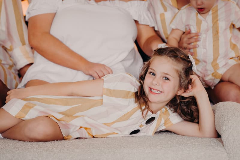A young girl in a striped dress lies on a carpeted floor smiling, surrounded by adults and children wearing matching striped outfits.