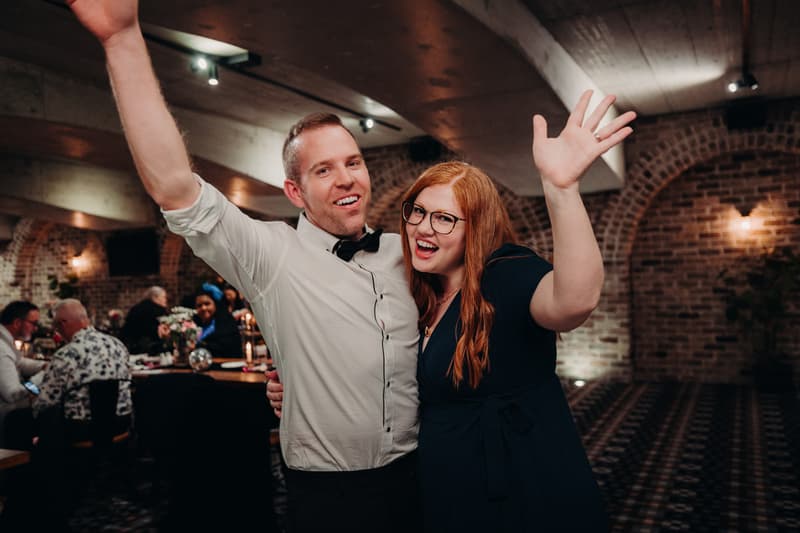 A man in a white shirt and black bow tie and a woman with long red hair and glasses pose with raised hands at the Sandstone Point Hotel — Cellar during the wedding reception.