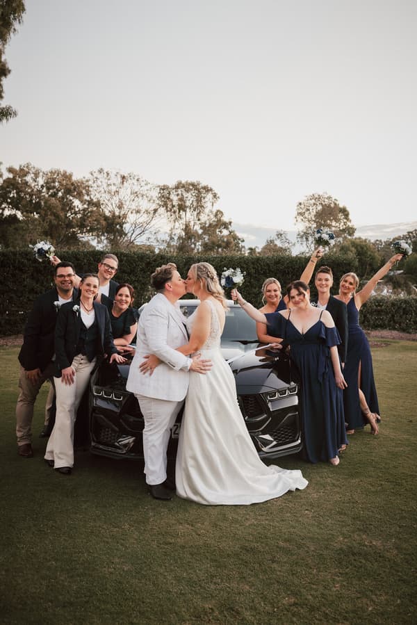 Mandy and Selina kiss in front of a black car at Sandstone Point Hotel, surrounded by their bridal party holding bouquets and smiling.