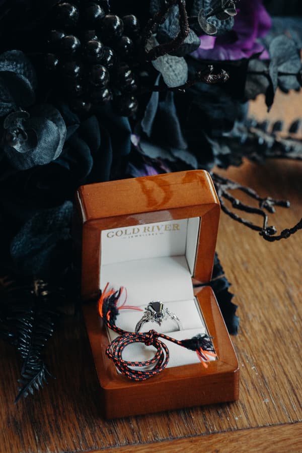 Close-up of a wooden ring box from Cold River containing a silver ring with a dark gemstone and a braided black and red cord, placed on a wooden surface with dark floral decorations nearby.