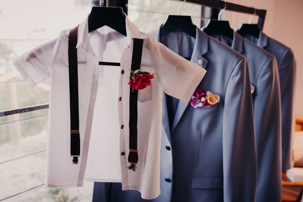 Groom's and groomsmen's attire hanging on a rack at Sandstone Point Hotel, including a white shirt with suspenders and a red boutonniere, and blue suits with floral boutonnieres.