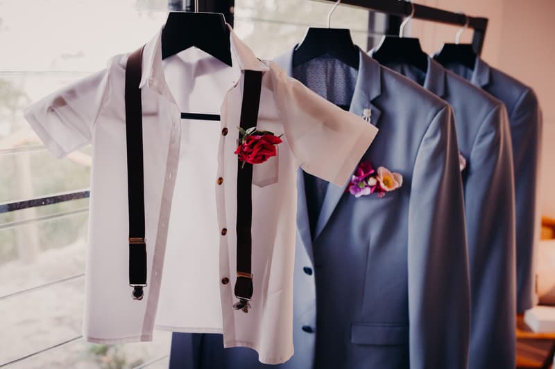 Groom's and groomsmen's attire hanging on a rack at Sandstone Point Hotel, including a white shirt with suspenders and a red boutonniere, and blue suits with floral boutonnieres.