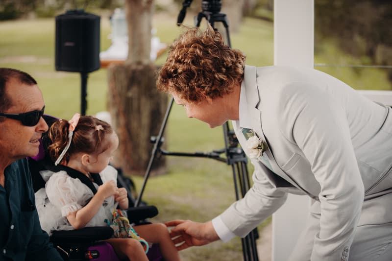 The groom Brodie, dressed in a light grey suit with a boutonniere, bends down to interact with a young flower girl seated in a wheelchair, while an older male guest wearing sunglasses sits beside her at Sandstone Point Hotel — Pavilion.